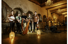 Download Foto: Eine Band mit Instrumenten steht in einem rustikalen Raum mit Backsteinwänden und Kronleuchtern. Bodenlampen leuchten.
