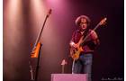 Download Foto: Ein Musiker spielt Bass auf einer Bühne mit rosa und violettem Licht. Ein weiteres Instrument steht auf einem Ständer daneben.