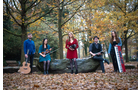 Download Foto: Fünf Musiker mit Instrumenten stehen im Herbstwald, umgeben von Laub und Bäumen.