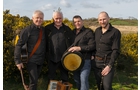 Download Foto: Vier Männer in schwarzer Kleidung posieren fröhlich im Freien, neben gelbem Ginster. Einer hält eine Bodhrán-Trommel.