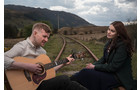 Download Foto: Ein Mann spielt Gitarre neben einer Frau. Beide sitzen auf Bahngleisen in einer ländlichen Landschaft.