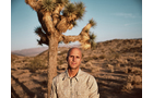 Download Foto: Ein Mann steht in einer Wüstenlandschaft neben einem Joshua-Tree. Er trägt ein helles Hemd, der Himmel ist klar.
