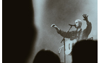 Download Foto: Eine Person mit Gitarre singt auf einer Bühne, die Arme ausgebreitet. Die Szene ist in stimmungsvollem, monochromem Licht gehalten.