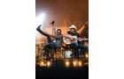 Download Foto: Zwei Musiker auf der Bühne, einer singt mit erhobenem Mikrofon, der andere spielt Gitarre. Bühnenlicht im Hintergrund.