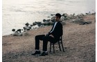 Download Foto: Ein Mann sitzt auf einem Stuhl am Kiesstrand, blickt aufs Wasser; im Hintergrund sind Steine und das Meer zu sehen.