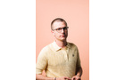 Download Foto: Ein Mann mit Brille in einem hellgelben Poloshirt steht vor einem pfirsichfarbenen Hintergrund.
