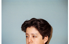 Download Foto: Eine Person mit dunklem Haar vor einem hellblauen Hintergrund schaut nach links aus dem Bild. Nur der Kopf ist sichtbar.