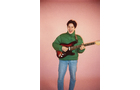 Download Foto: Person spielt E-Gitarre, trägt grünen Pullover und Jeans, steht vor rosa Hintergrund.
