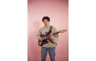 Download Foto: Person mit E-Gitarre vor rosa Hintergrund, trägt einen grauen Pullover und blaue Jeans, spielt konzentriert.