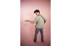 Download Foto: Eine Person in Jeans und Pullover spielt Gitarre vor einer rosa Wand. Sie steht seitlich und schaut über die Schulter zurück.