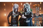 Download Foto: Zwei Personen in Glam-Rock-Kleidung posieren vor einer Bühne mit Schlagzeug und Lichtern. Beide haben auffällige Frisuren.