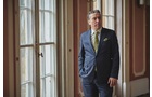 Download Foto: Ein Mann in Anzug und Krawatte steht in einem eleganten Raum mit großen Fenstern und verzierten Wänden.