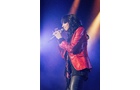 Download Foto: Eine Person singt auf einer Bühne, trägt eine rote Jacke und hält ein Mikrofon. Hintergrund mit blauem und goldenem Licht.