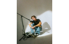 Download Foto: Eine Person mit Kopfhörern sitzt auf dem Boden eines hellen Raumes neben einem Mikrofonständer, entspannt und nachdenklich.