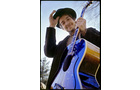 Download Foto: Mann mit Hut lächelt, hält eine Akustikgitarre. Blauer Himmel im Hintergrund, Bäume am Bildrand sichtbar.