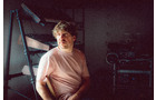 Download Foto: Ein Mensch in einem rosa T-Shirt sitzt in einem dunklen Raum mit Regalen und blickt nach links.