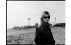 Download Foto: Eine Person mit Sonnenbrille steht am Strand, im Hintergrund sind ein Riesenrad und andere Strandattraktionen zu sehen.
