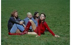Download Foto: Vier Personen sitzen auf einer Wiese, drei in blauen Kleidungsstücken, eine in rotem Kleid und roten Stiefeln, schauen ernst.