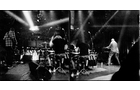 Download Foto: Schwarz-weiß Foto einer Band auf der Bühne, mit Schlagzeugern und Gitarristen, umgeben von Bühnenlicht und Publikum.