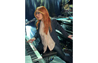 Download Foto: Eine Person mit langen roten Haaren spielt gleichzeitig auf zwei Keyboards auf einer Bühne. Sie trägt eine Brille und einen Blazer.