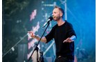 Download Foto: Ein Mann singt auf einer Bühne in ein Mikrofon, trägt einen schwarzen Pullover. Hinter ihm sind bunte Lichter und Nebel zu sehen.