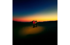 Download Foto: Zwei Personen stehen in einer dunklen Landschaft bei Sonnenuntergang, mit farbenfrohem Himmel von Orange zu Dunkelblau.