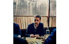Download Foto: Ein Mann sitzt an einem Tisch in einem Café, umgeben von Geschirr und einer Wasserflasche, vor einem Fenster mit Gardinen.