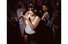 Download Foto: Eine Frau in weißem Top tanzt lächelnd in einem Club, umgeben von anderen feiernden Menschen. Die Beleuchtung ist schummrig.