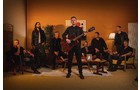 Download Foto: Gruppe von acht Personen in einem Raum, eine Person spielt Gitarre, andere sitzen oder stehen um sie herum, warme Beleuchtung.