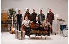 Download Foto: Eine Gruppe von acht Männern posiert in einem Studio mit diversen Musikinstrumenten, darunter Gitarren, ein Akkordeon und ein Schlagzeug.