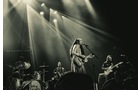 Download Foto: Eine Band spielt auf einer Bühne, umgeben von dramatischer Beleuchtung. Ein Sänger mit Gitarre steht zentral im Rampenlicht.