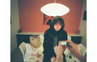 Download Foto: Eine Person mit dunklem Kapuzenpullover sitzt am Tisch, eine Hand fotografiert sie mit einer Kamera. Eine Lampe leuchtet hell darüber.