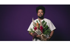 Download Foto: Eine Person mit Afrofrisur hält einen Strauß bunter Blumen vor einem dunkellila Hintergrund. Sie trägt ein weißes T-Shirt.