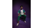 Download Foto: Eine Person in grüner Kleidung und weißen Sneakers sitzt auf einem Hocker vor einem lila Hintergrund.