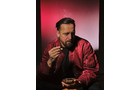 Download Foto: Ein Mann sitzt in roter Jacke vor rotem Hintergrund, hält eine Zigarre und ein Glas, schaut nachdenklich zur Seite.