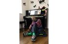 Download Foto: Person sitzt auf dem Boden vor einem Klavier, trägt Rollschuhe und bunte Kleidung. Gestickte Tiermotive sind an der Wand.