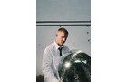 Download Foto: Ein Mann in einem weißen Kittel hält eine silberne Discokugel. Hintergrund ist eine graue Wand mit Metallrohren.