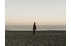 Download Foto: Eine Person steht allein auf einem kiesigen Strand bei ruhiger See im Hintergrund, während der Himmel pastellfarben leuchtet.
