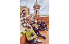 Download Foto: Eine Gruppe von Menschen sitzt lächelnd auf einer Terrasse mit einem verzierten Turm und bewölktem Himmel im Hintergrund.