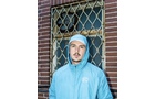 Download Foto: Ein Mann in einer hellblauen Kapuzenjacke steht vor einem vergitterten Fenster mit Backsteinmauerwerk.