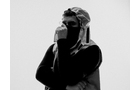 Download Foto: Person in Schwarz-Weiß, trägt eine Mütze und Maske, schaut zur Seite und hält die Hand vors Gesicht vor neutralem Hintergrund.