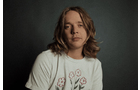 Download Foto: Person mit langen Haaren in einem weißen T-Shirt mit Blumenmotiv, vor einem dunklen Hintergrund.