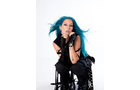Download Foto: Eine Person mit langen blauen Haaren und schwarzer Kleidung sitzt auf einem Stuhl vor weißem Hintergrund und schaut direkt in die Kamera.