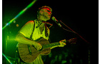 Download Foto: Eine Person spielt Gitarre und singt auf einer Bühne, im grünen Lichtschein. Sie trägt ein Mikrofon und ist in traditioneller Tracht gekleidet.