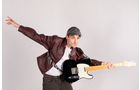 Download Foto: Person in Lederjacke und Mütze posiert energisch mit einer E-Gitarre, eine Hand ausgestreckt, vor grauem Hintergrund.