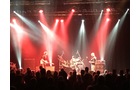 Download Foto: Eine Band auf einer Bühne, beleuchtet von roten und weißen Scheinwerfern, spielt vor einem Publikum.