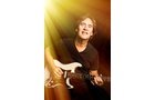 Download Foto: Eine Person spielt E-Gitarre im Rampenlicht, umgeben von warmem, goldenen Licht. Sie trägt ein schwarzes T-Shirt und lächelt.