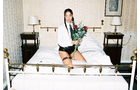 Download Foto: Eine Person sitzt auf einem Bett in einem Schlafzimmer, hält einen Blumenstrauß und trägt moderne Kleidung mit schwarzen Stiefeln.