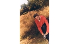 Download Foto: Eine Person in einem roten Pullover schnürt ihre Schuhe auf einem Feldweg, umgeben von hohen, goldenen Gräsern und Bäumen.