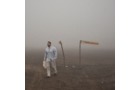 Download Foto: Ein Mann in weißer Kleidung geht durch neblige Landschaft, trägt eine Tasche. Im Hintergrund sind ein Schild und Stangen zu sehen.